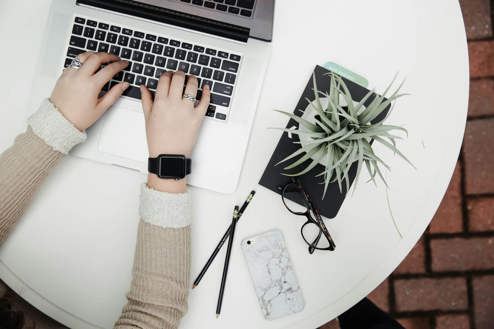 Hands typing on a laptop at a white desk with a plant and reading glasses